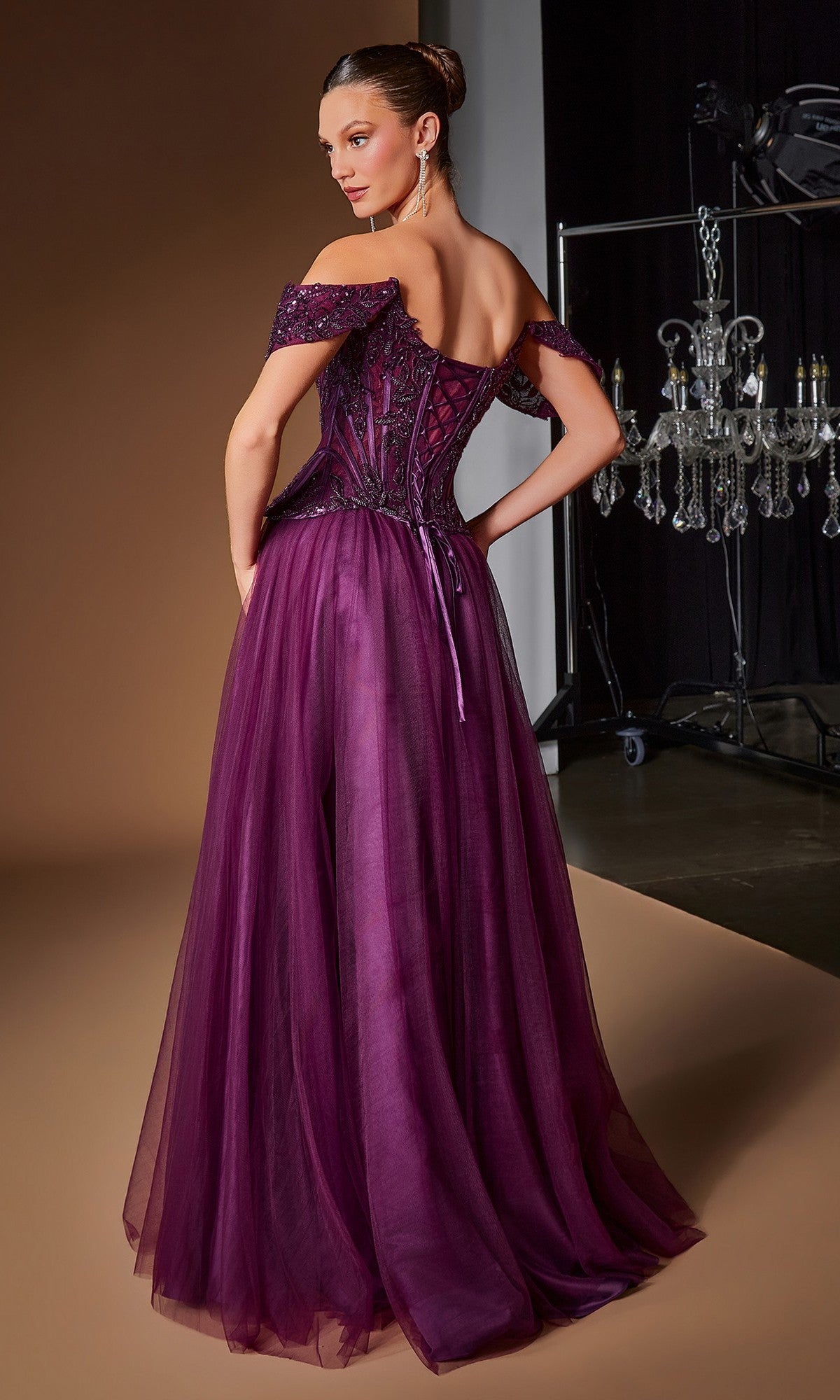 A woman stands with her back to the camera in a studio, modeling the Ladivine CD657—a floor-length, off-the-shoulder purple prom ball gown with a fitted corset bodice and sheer tulle skirt.